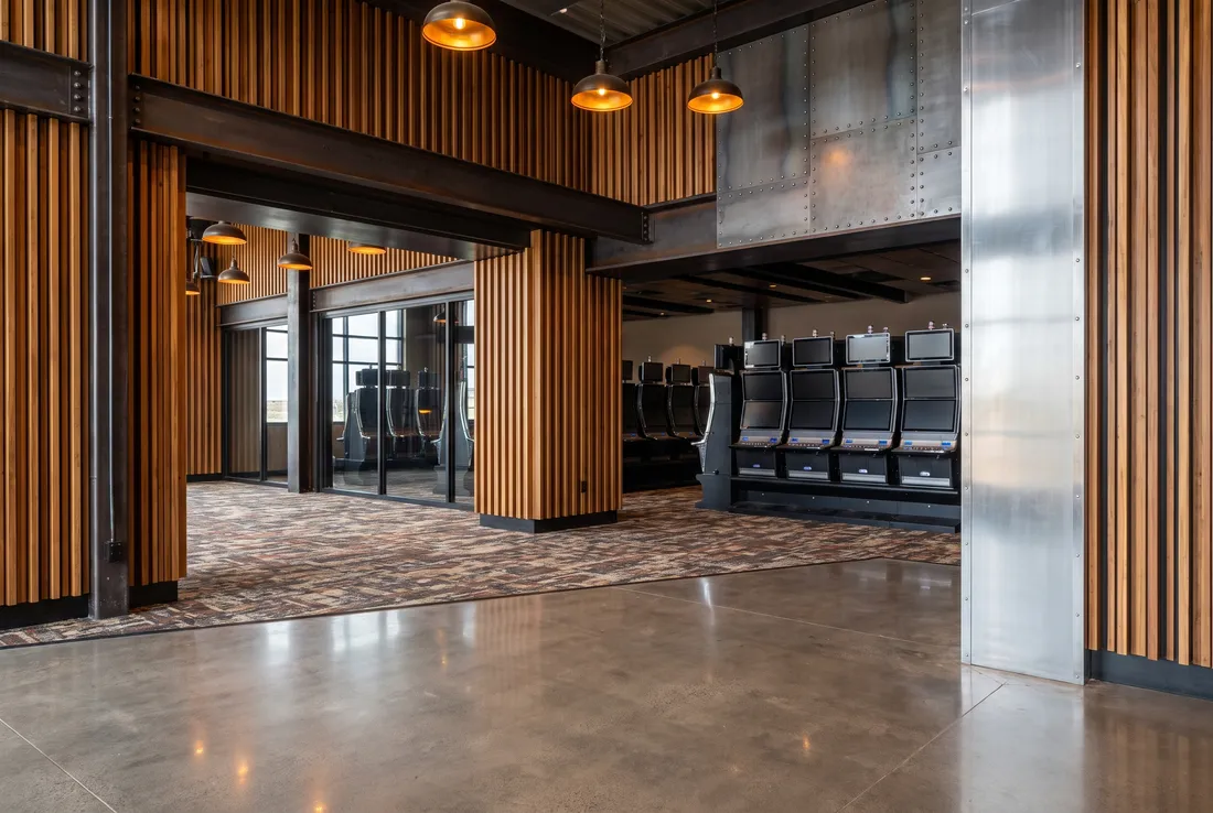 An entrance hall with vertical wood slats, dark steel beams, riveted metal panels and glass, with rows of slot machines with blank screens beyond.